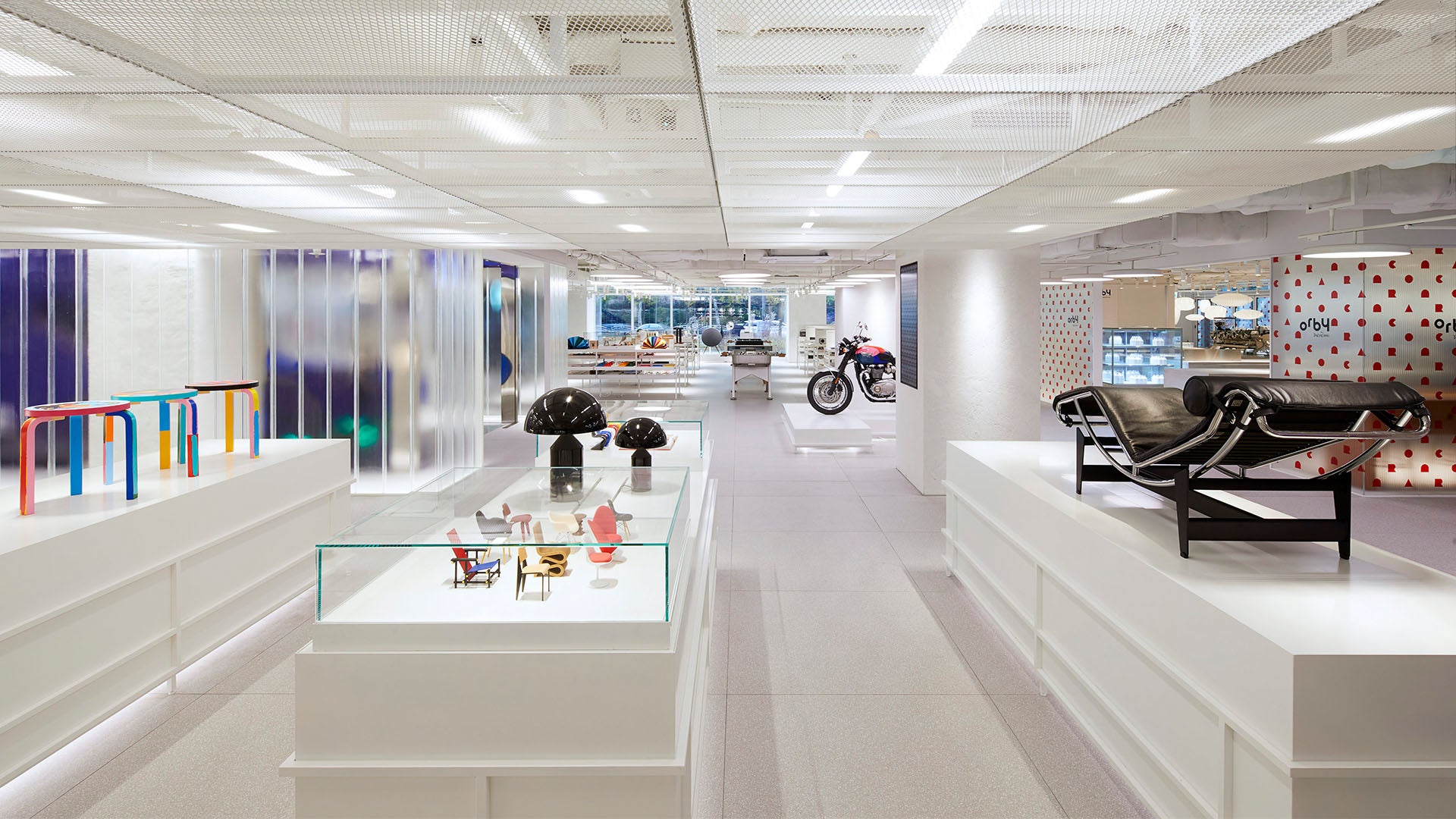 Inside The Conran Shop in Gangnam, a sleek, modern store with white displays and color-accented furniture. A motorcycle is showcased in the well-lit, expansive space featuring translucent panels and a variety of designer products. A black leather chaise lounge takes center stage in the foreground.